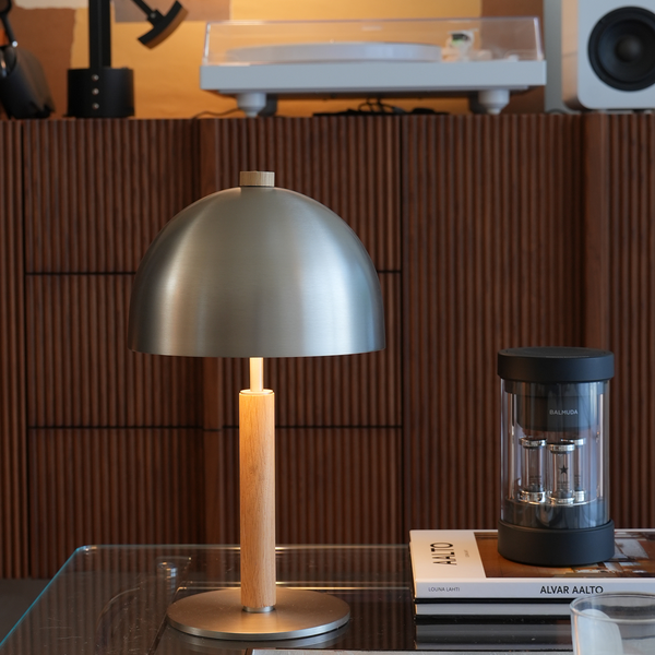 Table lamp with wooden base and metallic shade on a glass table, with books and a coffee grinder in the background.
