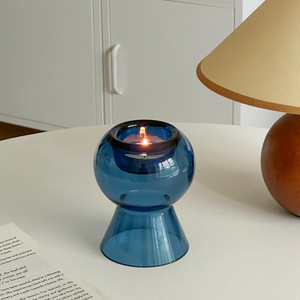 Blue glass candle holder with a lit candle on a white surface next to a book and lamp.