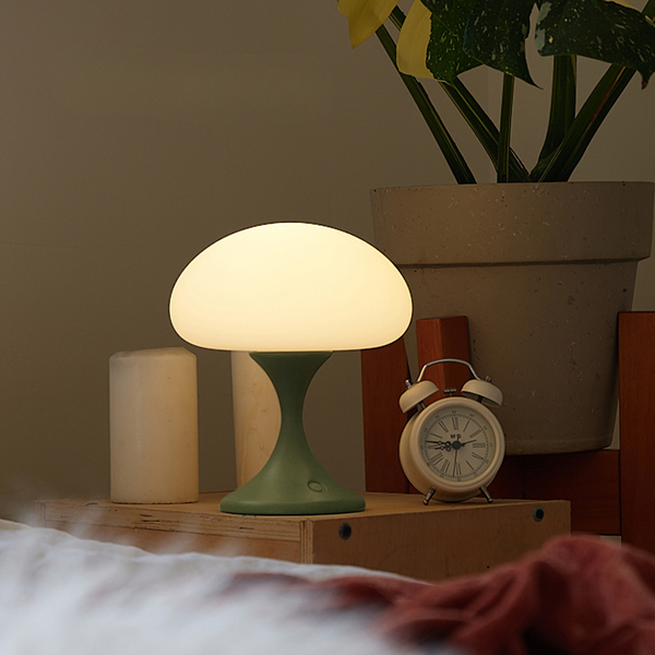 Mushroom-shaped lamp on a bedside table with a plant and alarm clock in the background.