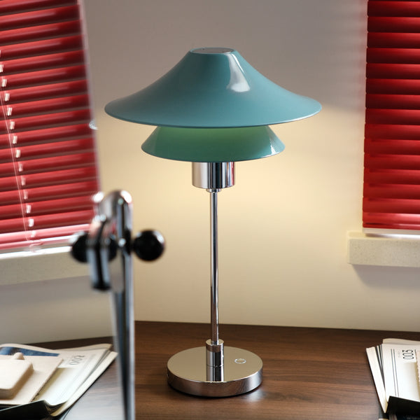 Table lamp with a green shade on a desk in a room with red blinds.