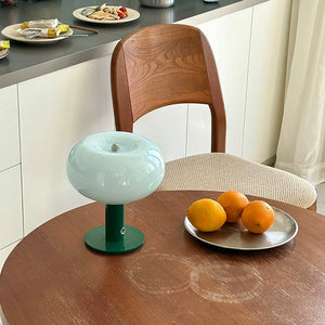 Round wooden table with a small green lamp and plate of oranges in a kitchen setting.