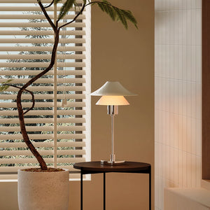 Modern table lamp on a small round table next to a potted plant with blinds in the background.