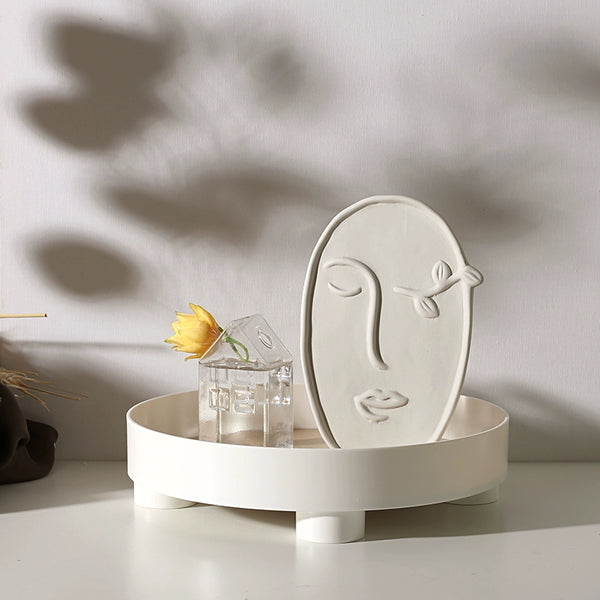 Decorative white face-shaped object on a round stand with a yellow flower and small glass house in the background.